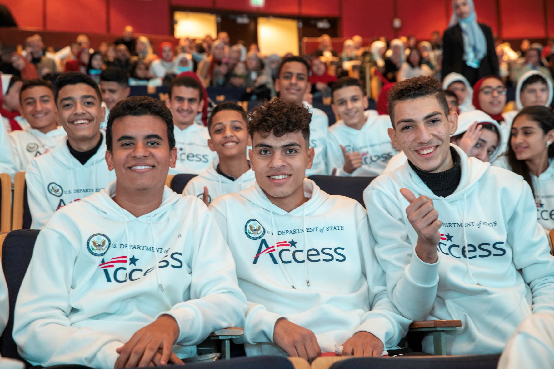 Boys sitting in a hall full of young people. Text on their t-shirts:Access, U.S. Department of State