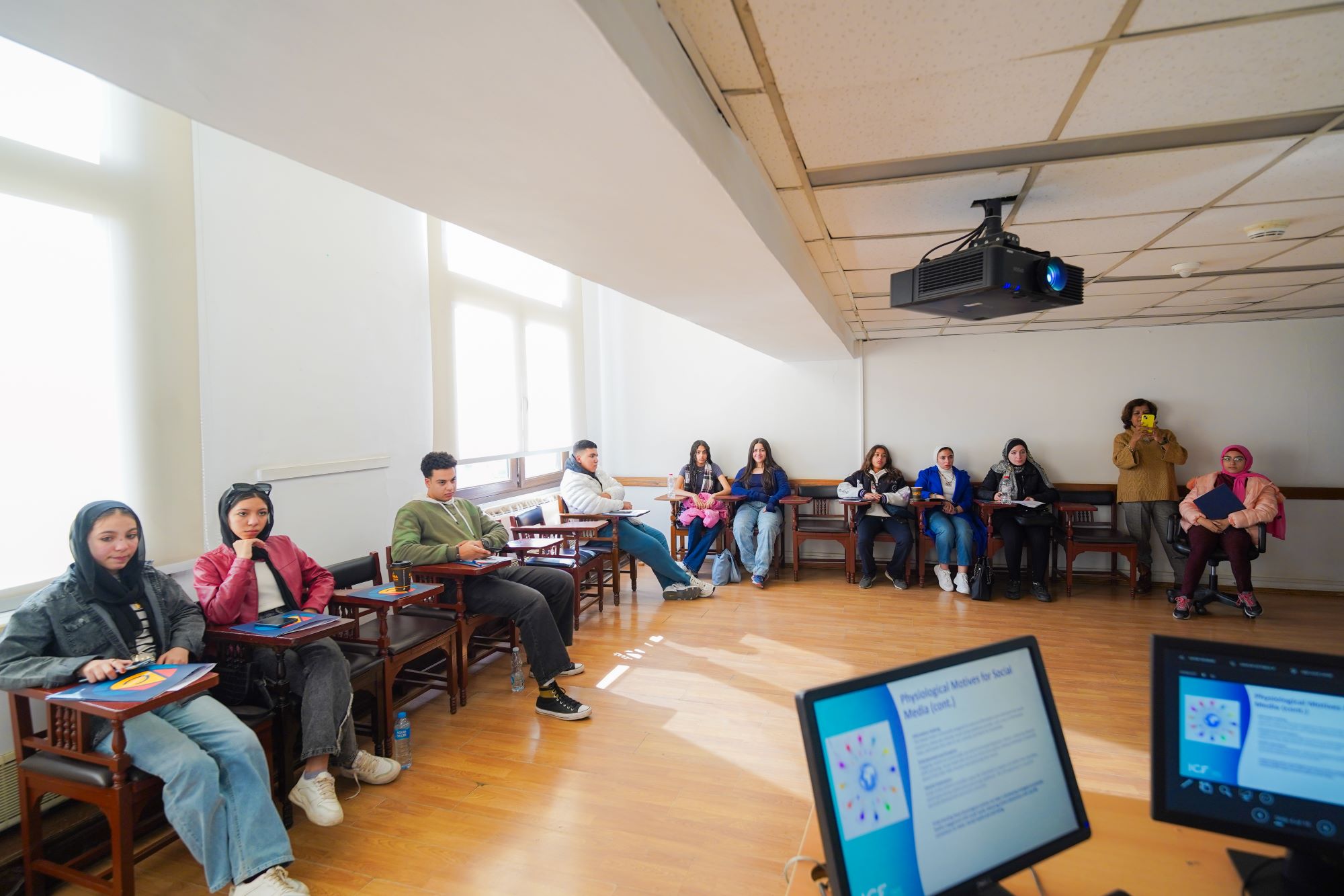 A group of students sitting in a semi-circle of wooden tablet-arm chairs, listening to a presentation with computer monitors visible in the foreground.