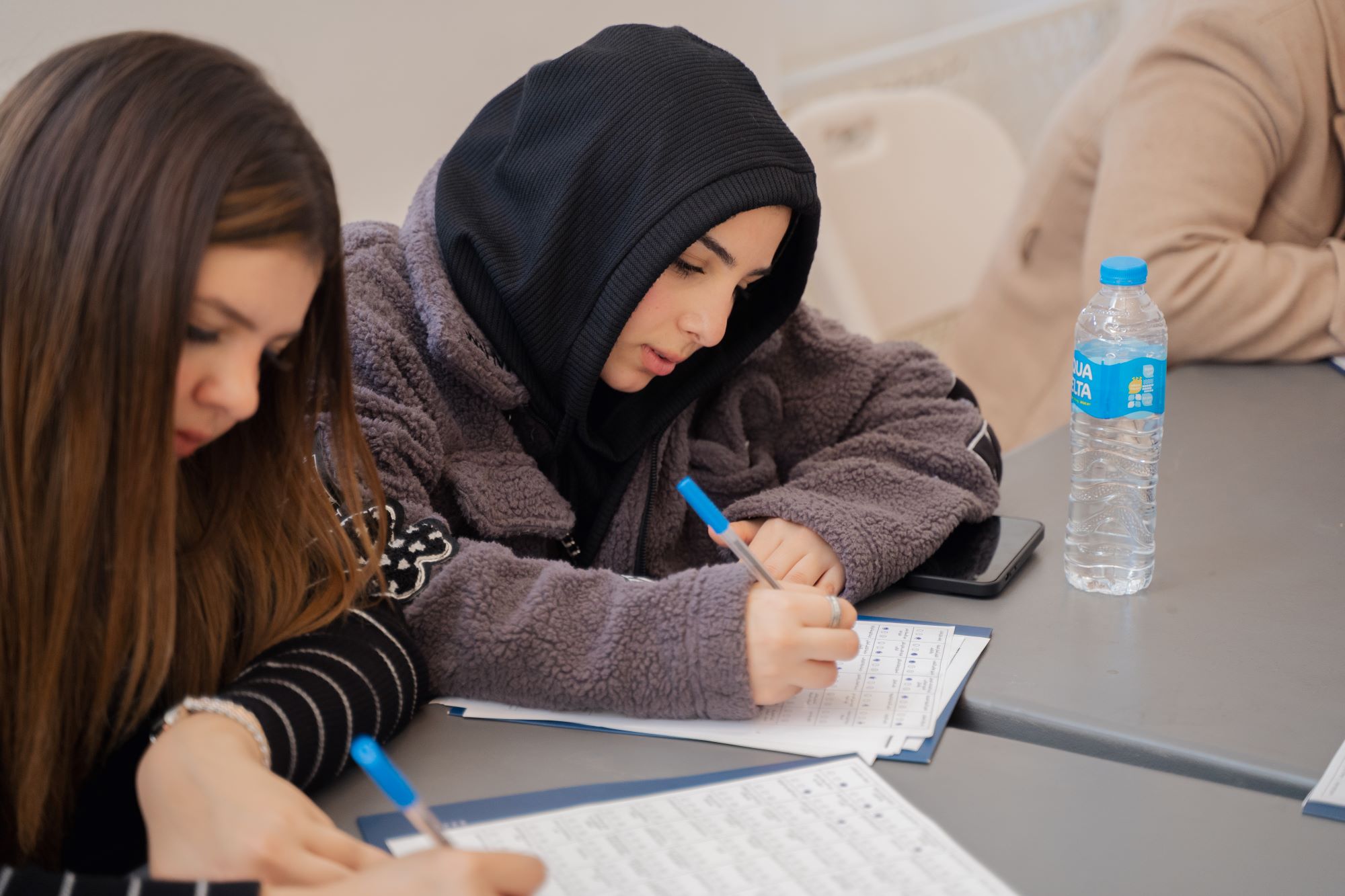 Two young women sitting at a table side-by-side, focused on writing and filling out test papers.