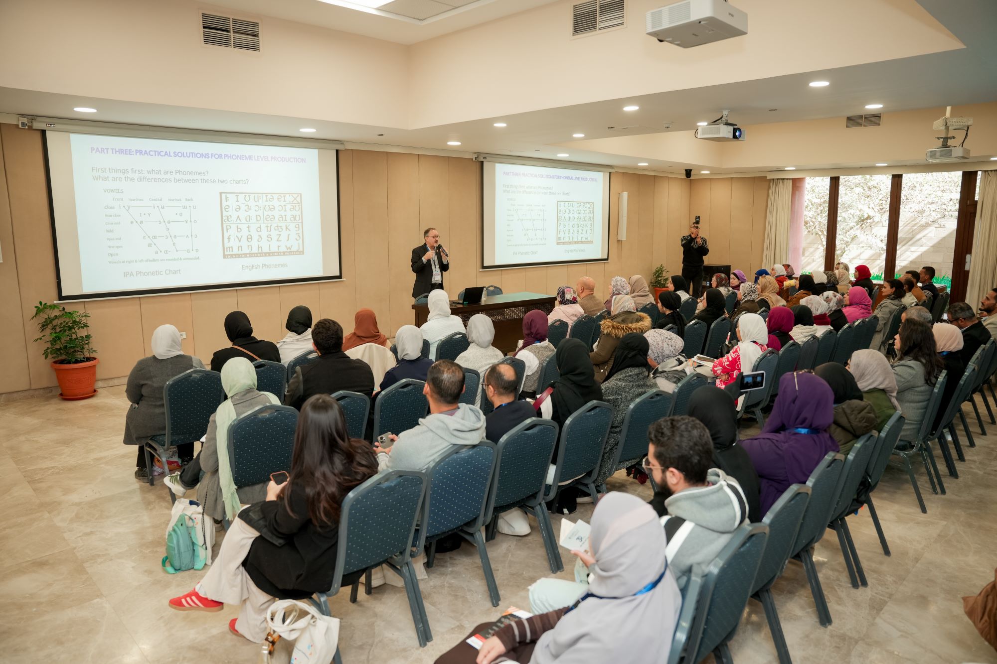 A speaker stands at the front of a crowded lecture hall, presenting slides on phonetics to a seated audience.