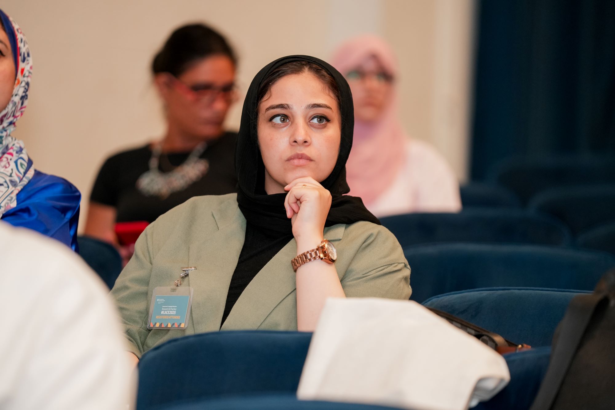 A woman wearing a hijab and an olive-green blazer sits attentively in an auditorium, listening with her hand resting on her chin.