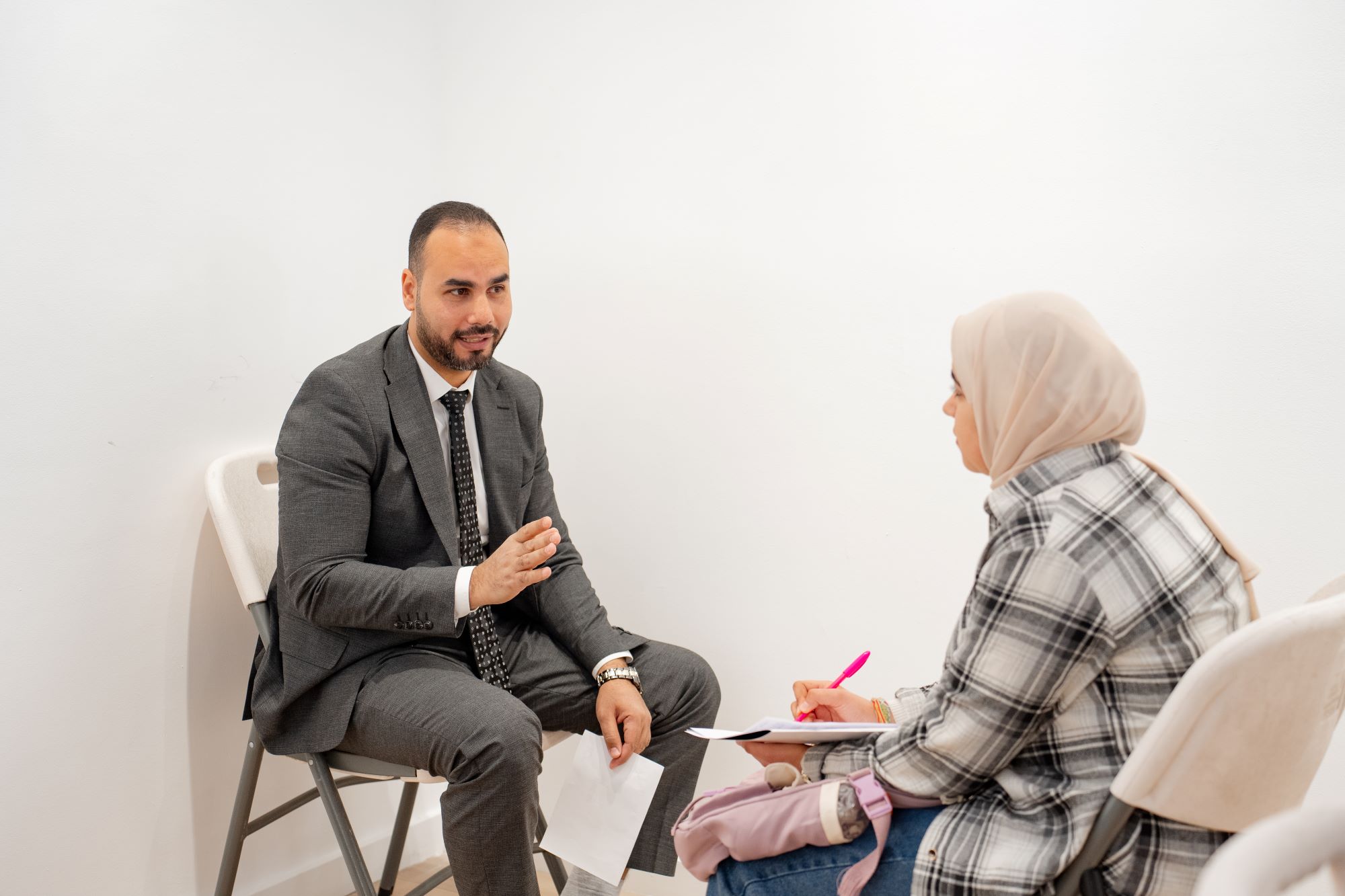 A man in a grey suit gestures while speaking to a woman wearing a hijab who is taking notes.