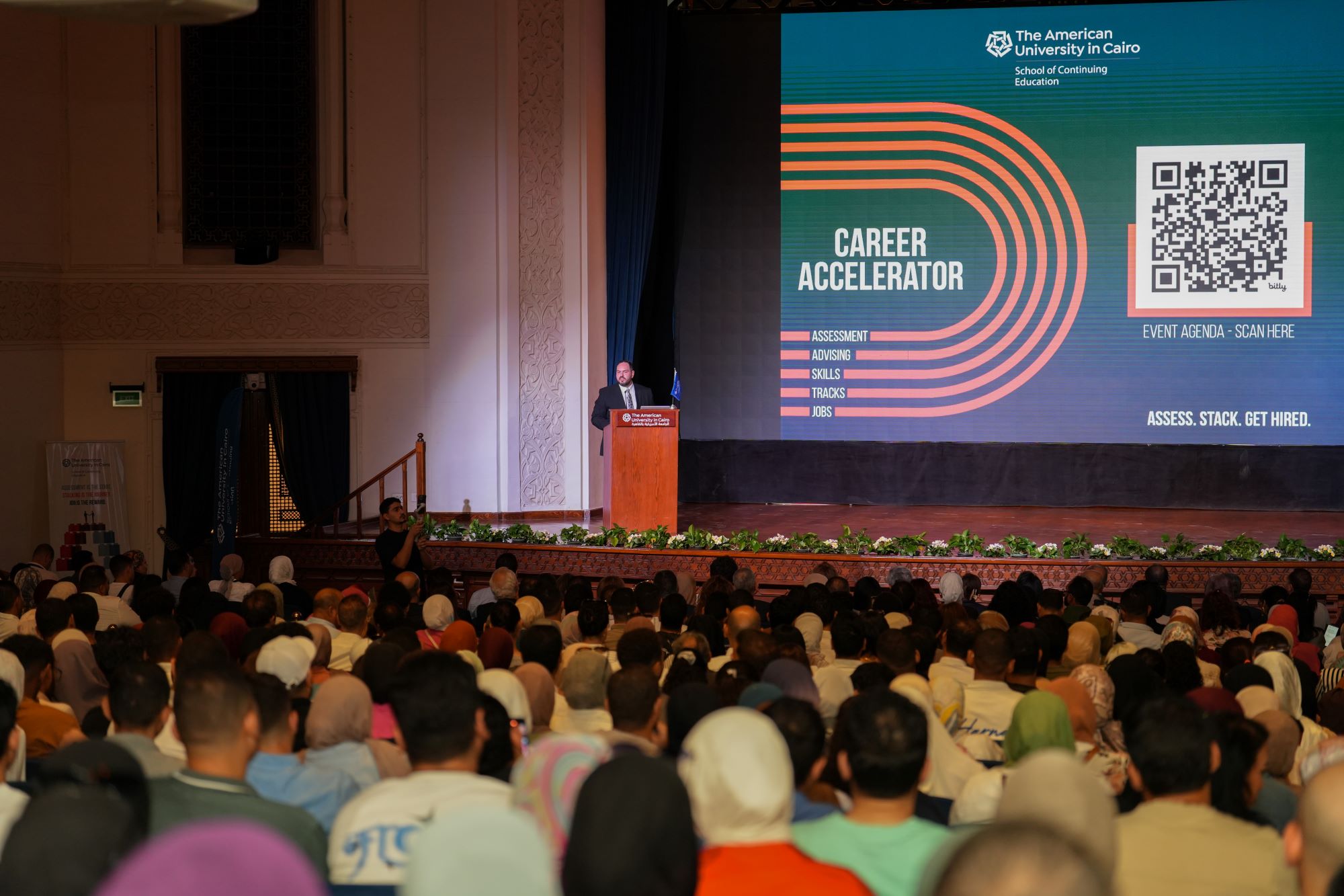 A grand, packed auditorium hosting the "Career Accelerator" keynote session by The American University in Cairo, featuring a presenter on stage addressing a massive audience.
