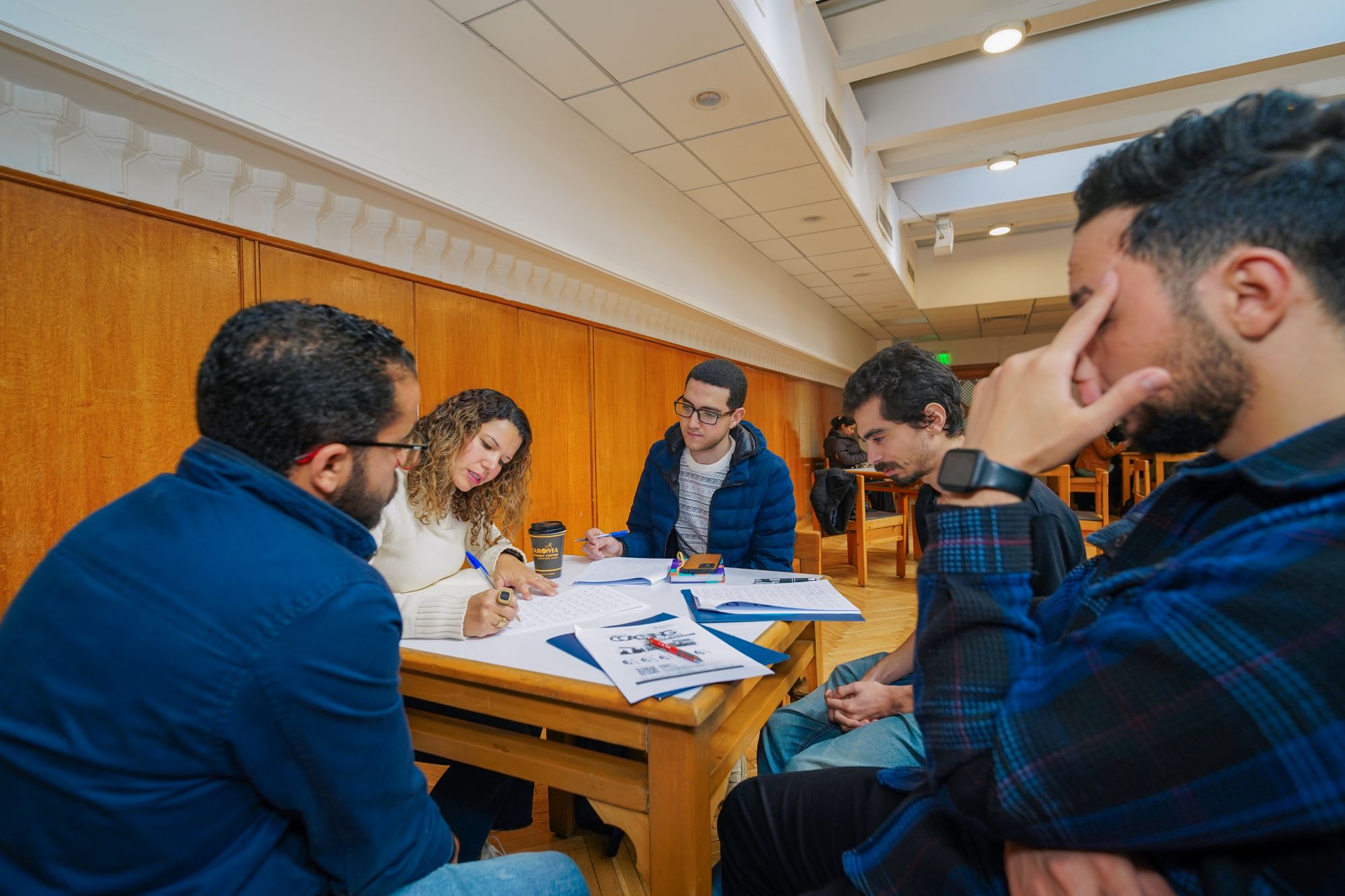 A group of young adults sitting around a wooden table, collaborating and writing on papers during a workshop.
