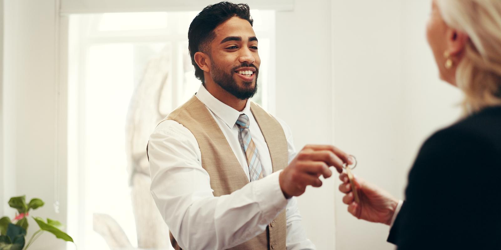 Happy man, keys and hotel lobby for businesswoman, welcome or registration for accommodation. People, check in and concierge service with client, help desk and hospitality for suite booking at lodge