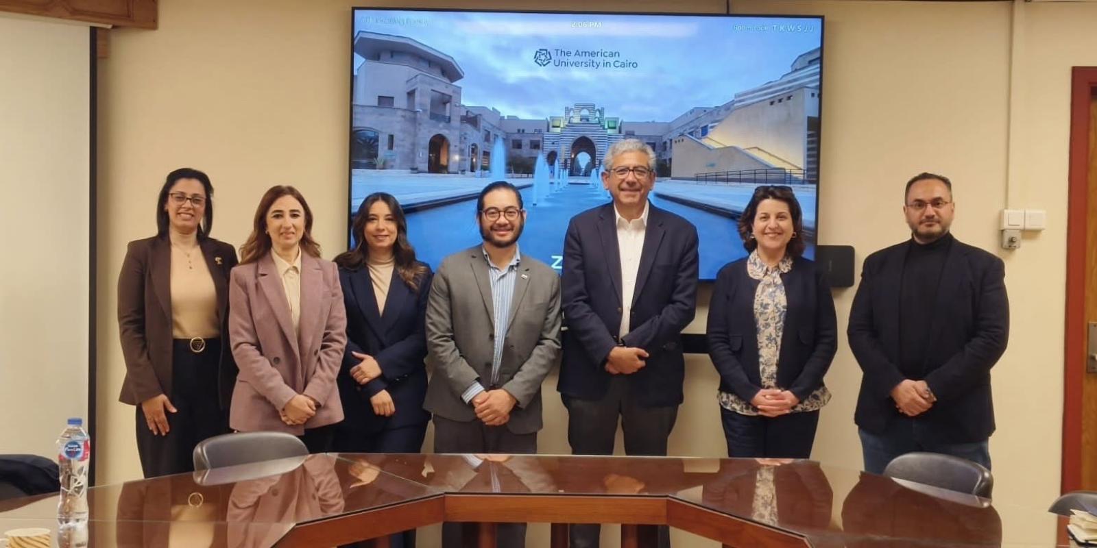 A professional group of men and women pose together in front of a digital screen displaying a photo of The American University in Cairo campus.