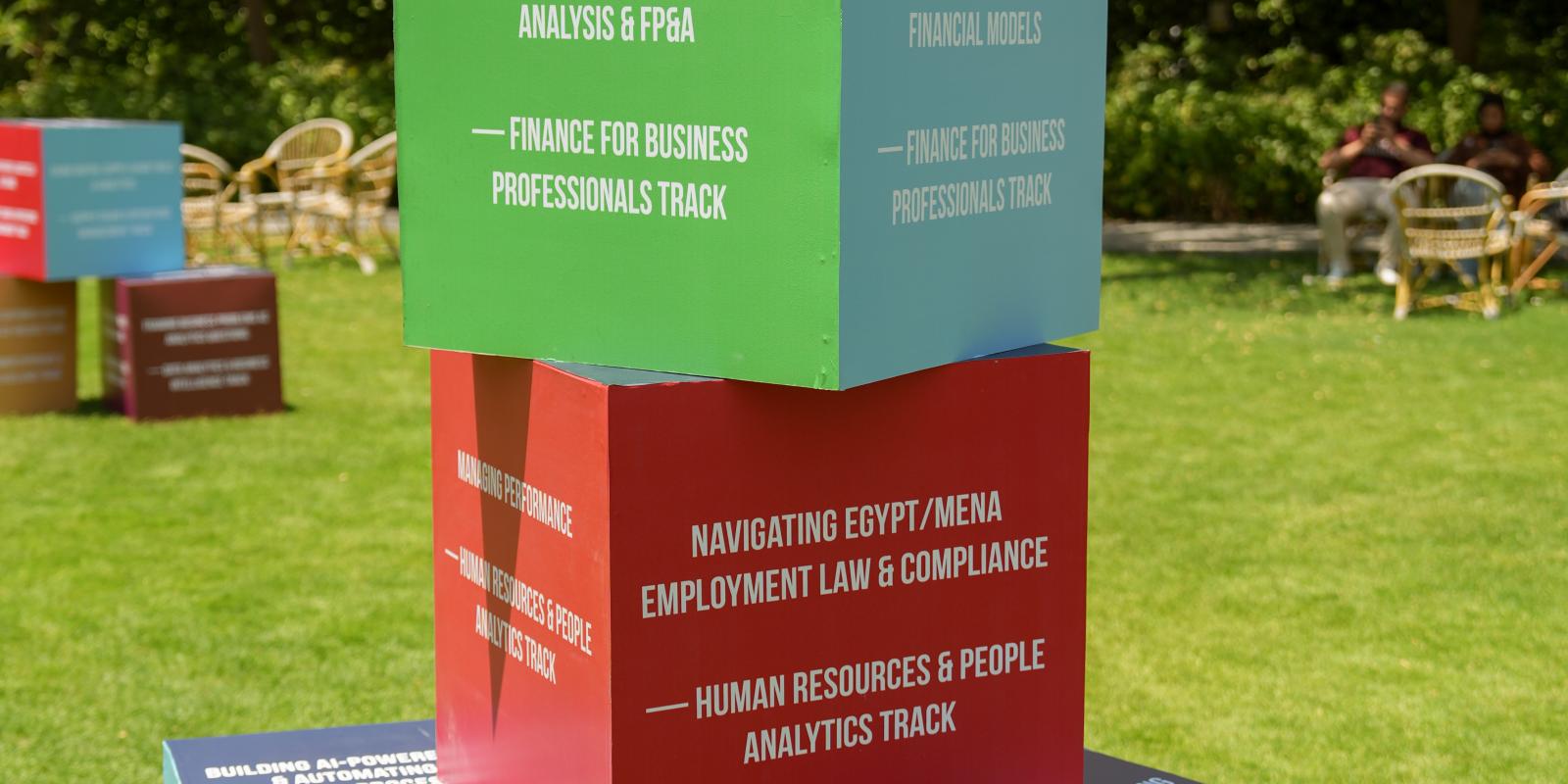 A detailed close-up shot of a colorful, stacked cube installation displayed on a sunlit campus lawn, highlighting specific educational modules and professional development tracks.