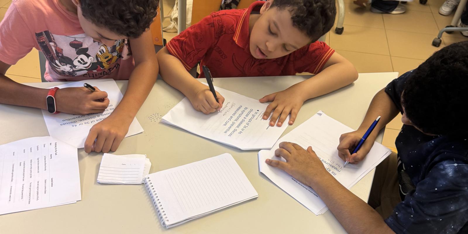 Three boys sit closely around a modular classroom desk, deeply focused on writing on separate sheets of paper.