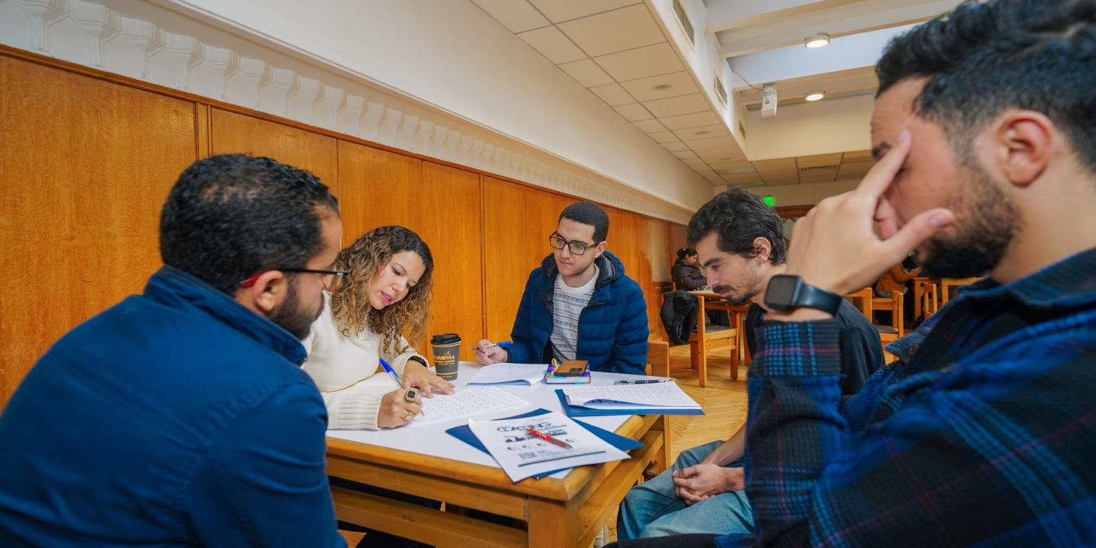 A group of young adults sitting around a wooden table, collaborating and writing on papers during a workshop.