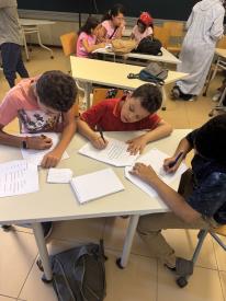 Three boys sit closely around a modular classroom desk, deeply focused on writing on separate sheets of paper.